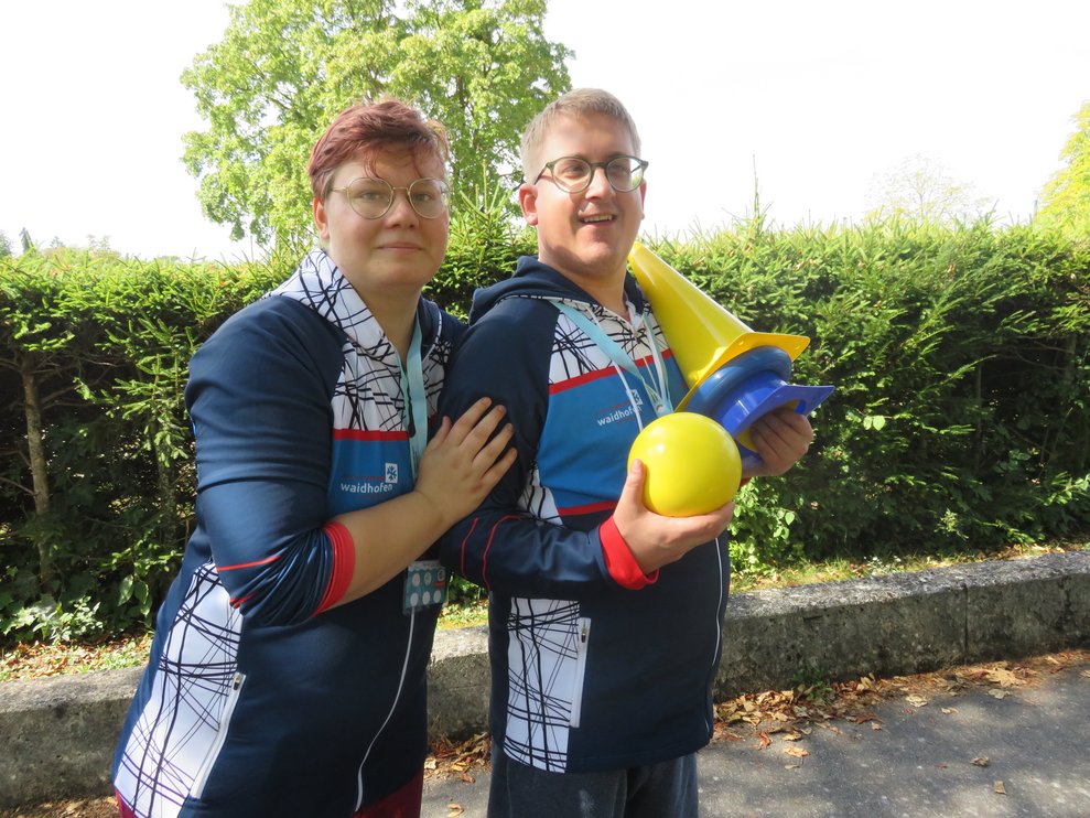 Eine Frau steht neben einem Mann, der Mann hält einen Ball und bunte Hütchen.