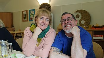 Zwei Personen sitzen am Tisch und lachen in die Kamera
